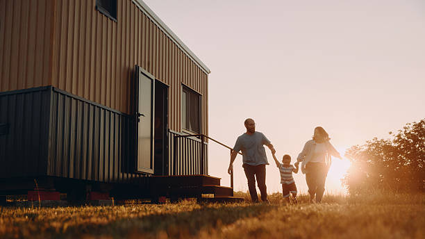 Family outdoor fun in front of a tiny house A warm outdoor family moment in the late afternoon sun in front of a modern tiny house on wheels. A mother, father, and their young son hold hands and run together across a sunlit lawn, capturing joyful, carefree play in a small-space living setting. The tiny house features wooden siding and a bright exterior, with golden hour lighting creating a cozy, welcoming atmosphere. the tiny house stock pictures, royalty-free photos & images