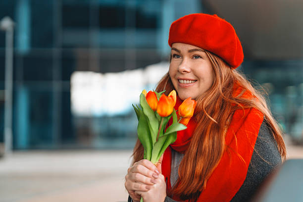 A cheerful young woman with long red hair, wearing a red beret and scarf, enjoys a sunny day while holding bright tulips in a lively city location.