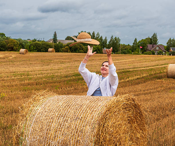 Happy woman standing behind hay bale and tossing her hat into the air in an open field. Scene full of movement, happiness, and carefree country freedom.