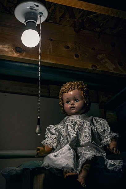 A creepy old baby doll sits on a high shelf in an attic by a lightbulb with a pull string.