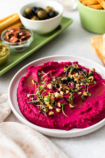 Beetroot hummus is placed in the center of a charcuterie board, surrounded by snacks like olives, nuts, cheese, and crispy breadsticks. A light, inviting setup showcases vibrant colors