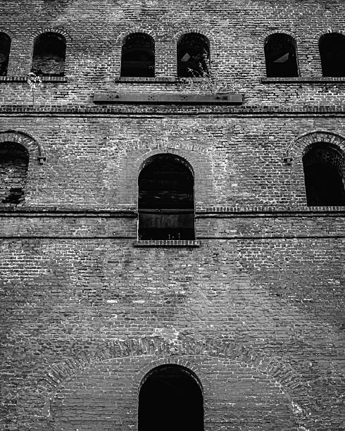 The facade is structured by a symmetrical arrangement of three levels with semicircular arched windows, most of which are empty and dark. The rough, coarse bricks dominate the texture, enhanced by deep shadows, which gives the image a harsh, industrial and timeless expression. This structure radiates a quiet, architectural strength, marked by the traces of time.