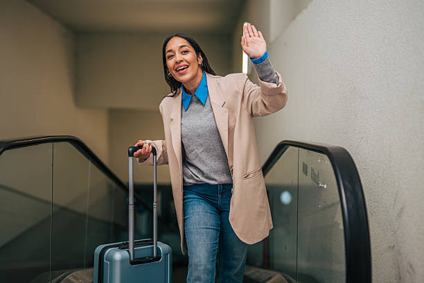 Smiling woman traveling on an escalator while holding a rolling suitcase and waving to people, representing greetings, farewells and embarking on a journey
