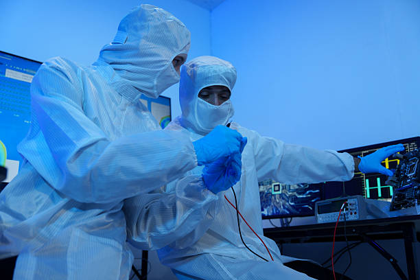 Engineers in sterile suits work at a semiconductor manufacturing plant using microscopes in a lab.