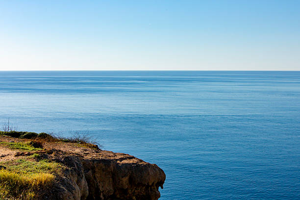 A peaceful coastal scene from Antalya, Turkey, showing a rocky cliff covered with green grass and wild plants. The cliff overlooks a calm blue sea that stretches to the horizon under a clear sky. The sunlight highlights the natural textures and colors, creating a relaxing and open atmosphere. This image is ideal for use in travel promotions, nature blogs, wellness content, vacation brochures, or websites related to tourism, mindfulness, and outdoor activities. It evokes feelings of calm, freedom, and connection with nature.