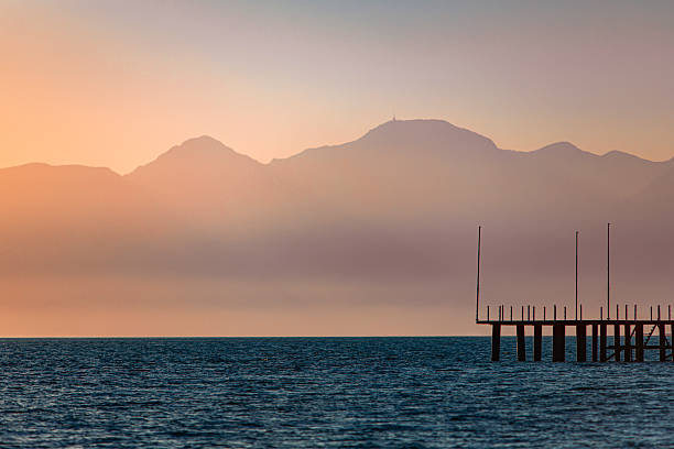 A tranquil seascape showing calm waters during sunset, with a silhouette of a pier extending into the sea on the right side. In the background, layers of mountains rise above the coastline, including the peaks above Antalya, softly lit in warm tones of orange and purple. The tallest mountain features a small structure or antenna at its summit. The scene is peaceful and atmospheric, ideal for use in travel content, nature blogs, wellness and meditation themes, vacation brochures, and materials related to coastal landscapes or sunset views.