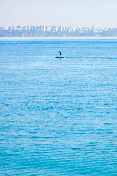 A peaceful seascape showing a lone paddleboarder gliding across calm blue waters. In the distance, the city of Antalya is visible along the coastline, with numerous high-rise buildings forming a soft urban silhouette. The clear sky and gentle haze near the horizon create a tranquil and spacious atmosphere. This image is suitable for travel and lifestyle content, wellness blogs, vacation brochures, and materials related to coastal recreation, solitude, or Mediterranean destinations.