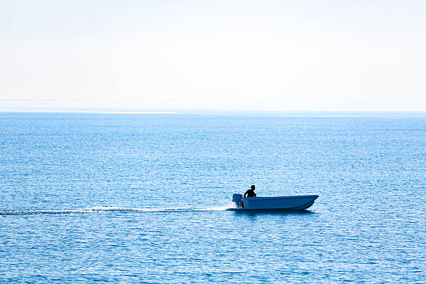 A small motorboat with a single person onboard travels across calm, vibrant blue waters under a clear sky. The boat creates a gentle wake as it moves forward, emphasizing motion and solitude. The wide expanse of sea and absence of other boats or land convey a peaceful, minimalist atmosphere. This image is suitable for travel and lifestyle content, marine recreation promotions, wellness blogs, vacation brochures, and materials related to freedom, exploration, or coastal living.