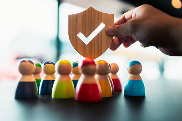 istock Hand holding wooden shield with check mark protecting diverse wooden people figures, symbolizing group protection, insurance, teamwork safety, and community care. 2242202619