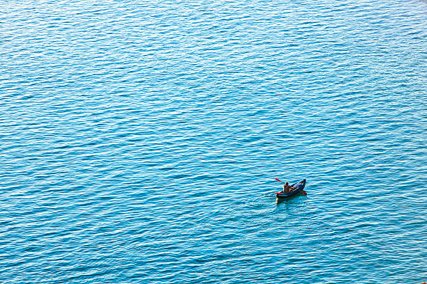 A single kayak floats peacefully on a vast expanse of calm blue water. The paddler is seated alone, with a red flag attached to the kayak, creating a focal point in the minimalist scene. Gentle ripples surround the vessel, and the clear sky enhances the tranquil atmosphere. This image is suitable for travel and lifestyle content, wellness blogs, vacation brochures, and materials related to solitude, marine recreation, or coastal exploration.