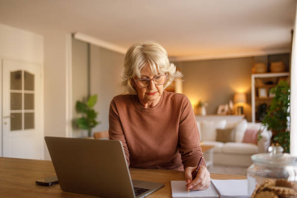 istock Elderly woman writing expenses in notepad and using laptop at desk while managing budget in living room 2242212320