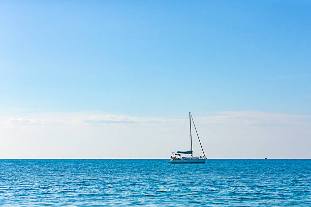 A serene maritime scene featuring a sailboat floating on calm, expansive blue waters beneath a clear sky. The boat has a white hull and a blue cockpit cover, with its sails down and mast upright, suggesting it is anchored or drifting. The horizon line separates the tranquil sea from the sky, which contains a few wispy clouds. This image is suitable for travel and lifestyle content, sailing and marine recreation promotions, wellness blogs, vacation brochures, and materials related to freedom, exploration, or coastal relaxation.