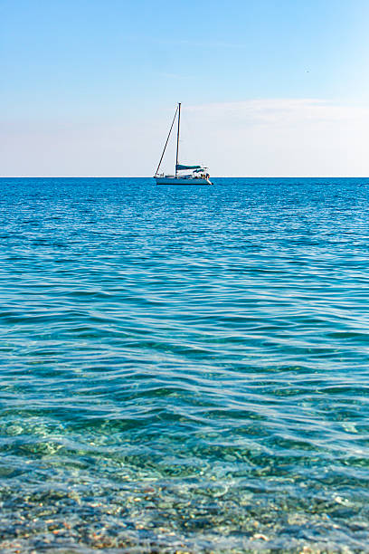 A serene maritime scene featuring a sailboat floating on calm, expansive blue waters beneath a clear sky. The boat has a white hull and a blue cockpit cover, with its sails down and mast upright, suggesting it is anchored or drifting. The horizon line separates the tranquil sea from the sky, which contains a few wispy clouds. This image is suitable for travel and lifestyle content, sailing and marine recreation promotions, wellness blogs, vacation brochures, and materials related to freedom, exploration, or coastal relaxation.