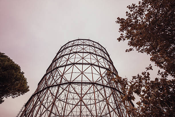 Low-angle view of a steel gasometer structure rising against a cloudy sky. The geometric framework and metallic symmetry highlight the industrial heritage and architectural design of urban infrastructure.