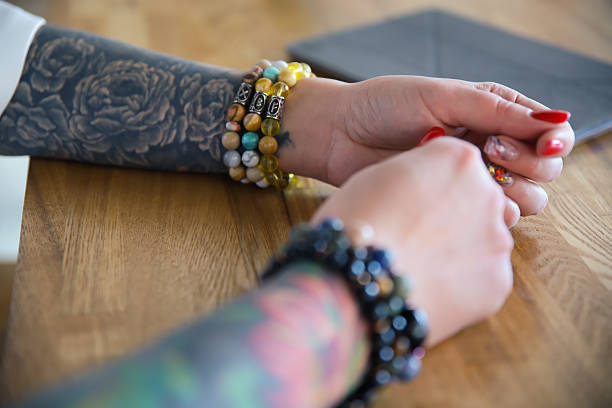 Stacked gemstone bracelets on tattooed wrists by a coffee table, colorful beads and ink