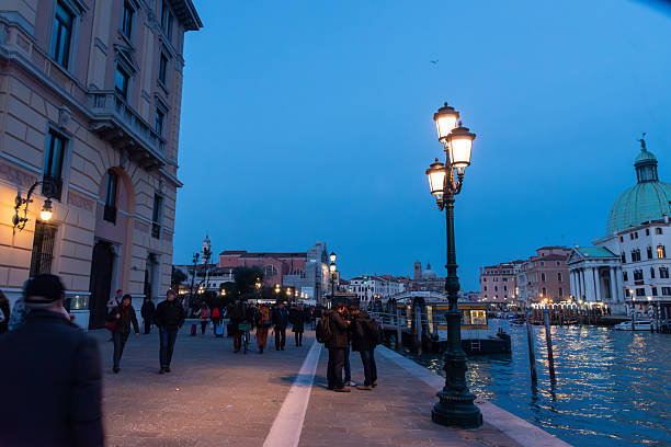 Tourist is standing on a wooden pier in Venice with gondolas moored next to it, illuminated by a lamppost at dusk, creating a charming travel scene