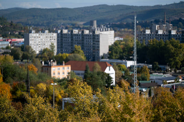 Urban landscape featuring residential apartment blocks surrounded by colorful autumn trees with shades of green, yellow, and orange. In the background, forested hills rise under a clear blue sky, creating a contrast between nature and urban architecture. The warm light adds a peaceful and balanced atmosphere to the scene.