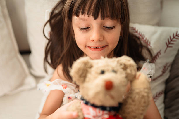 Adorable young girl smiling while holding a teddy bear in her hands on a cozy sofa. Soft daylight and warm tones capture a peaceful, heartwarming childhood moment at home.