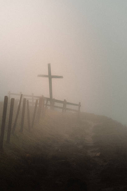 Cross in thick fog creates a mystical atmosphere. The photo shows a wooden cross standing on a foggy hillside beside a rustic fence. The thick mist softens the scene, creating a mysterious and almost spiritual atmosphere. The dim light and muted colors give the image a somber, contemplative mood.