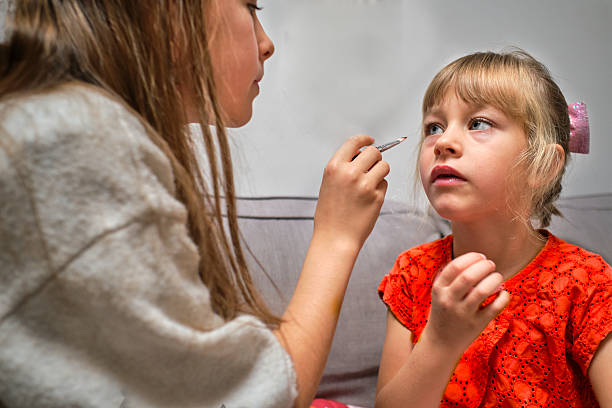 An older sister carefully applies makeup to her younger sister, who sits patiently on the couch in a bright orange dress. The moment feels playful yet calm, showing the bond between siblings during an everyday home activity. Natural light and soft tones highlight their concentration and affection, creating a warm and authentic family scene.