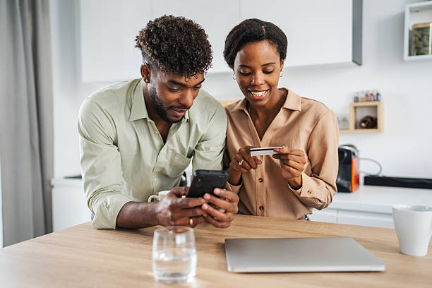 Young black couple shopping online in their modern kitchen, smiling as they browse on a smart phone and make a secure credit card payment together at home