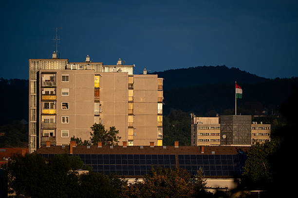A residential apartment block illuminated by warm sunset light stands behind a row of rooftops with solar panels. A Hungarian flag waves in the distance against a backdrop of green hills and deep blue sky, symbolizing sustainability and urban development in harmony with nature.