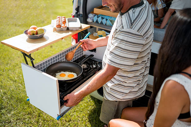 A Caucasian man prepares eggs in a skillet on a portable stove during a sunny outdoor camping trip, while a family enjoys the relaxing morning together.
