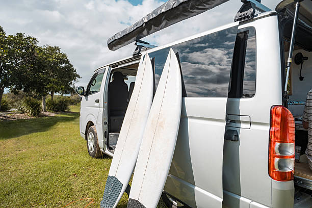 A silver camper van with two white surfboards leaning against it, parked on green grass under a partly cloudy blue sky near trees.