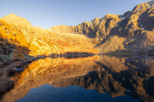 Golden autumn trees and rugged mountain peaks reflected in a calm lake at sunset. Scenic mountain landscape with clear water and warm sunlight. Nature and outdoor scenery.
