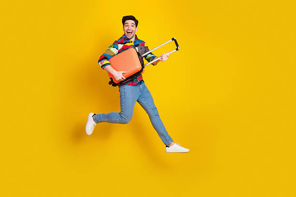istock Young man joyfully jumps with orange suitcase against a bright yellow background 2242323291