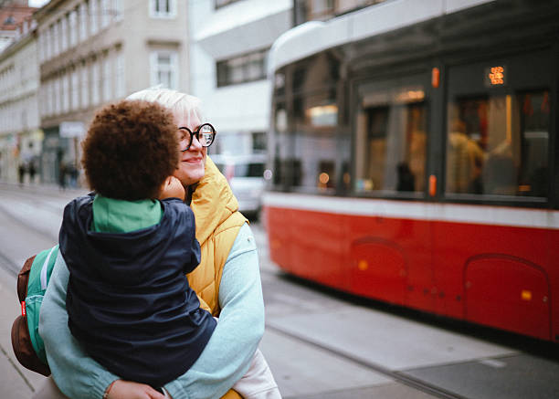 A loving mother holds her 3-year-old son while waiting for the tram at a city station. The toddler faces away from the camera and plays with his mother’s earring, as she smiles peacefully with her eyes closed. A tram passes by in the background, creating a beautiful mix of everyday urban life and heartfelt family connection. Perfect for themes of motherhood, parenting, affection, city life, transportation, and modern family moments.