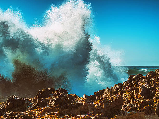 Violent ocean wave explodes dramatically over jagged, reddish-brown rocks, under a clear blue sky