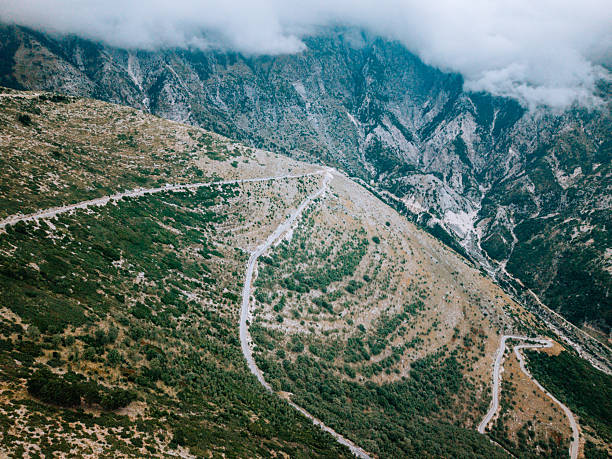 Aerial view of a winding mountain road cutting through rocky terrain with scattered vegetation. Misty clouds hover over steep mountain peaks. Scenic landscape with rugged natural beauty.