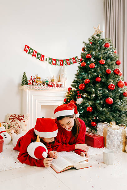 Asian mother and daughter in Santa hats read a book by decorated Christmas tree and fireplace. Surrounded by festive decor, gifts, and warmth, the holiday scene captures family, children, and joy.