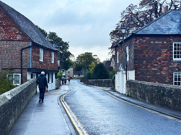 Looking over Harnham Bridge in Salisbury, England, on a cloudy day. The scene features a narrow, curving road with double yellow lines, flanked on both sides by low stone walls. Historic buildings with brick and timber-frame construction and tiled roofs line the street.