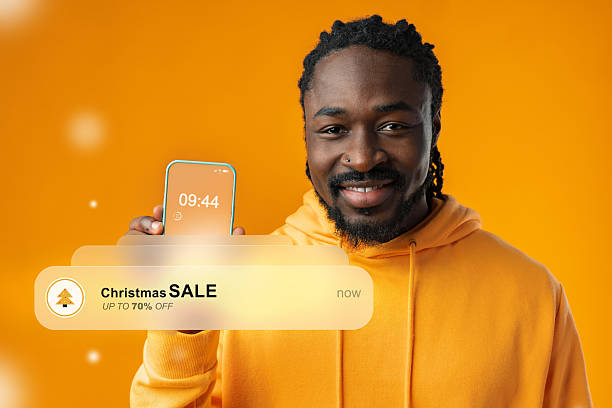 istock Man smiles while holding smartphone showing Christmas sale notification on bright orange background 2242336525
