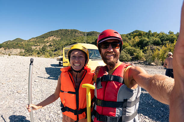 Happy man and woman wearing helmets and life jackets holding paddles on rocky riverbank. Outdoor adventure enthusiasts preparing for white water rafting trip. Excited friends enjoying nature and water sports.