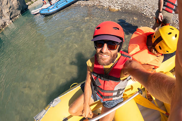 Bearded man wearing helmet and life jacket taking selfie on yellow inflatable raft in clear river water. Outdoor adventure with friends enjoying white water rafting. Group rafting activity on river.