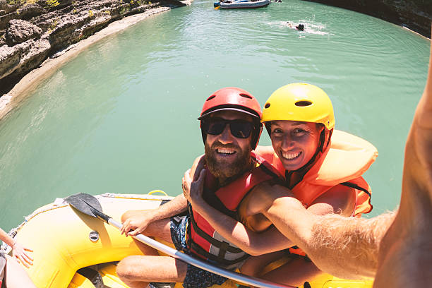 Smiling couple wearing helmets and life jackets on yellow raft in calm river water. Happy young adults enjoying outdoor water sport activity. Adventure and travel lifestyle with friends.