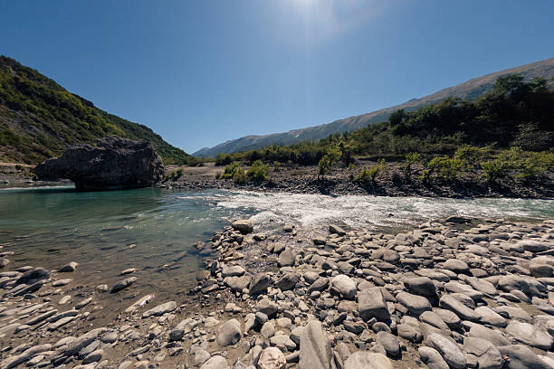 Clear river water flowing over rocks near a rocky riverbank with green hills and mountains under a bright blue sky. Scenic natural landscape with fresh water and outdoor wilderness. River nature scene.