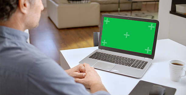 Mature Caucasian male working from home on a laptop with a green screen, featuring a modern home office setup. The individual appears focused and concentrated, highlighting a professional remote work environment.