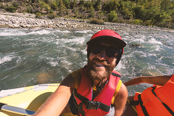 Bearded man wearing helmet and life jacket smiling on yellow raft in fast river. Outdoor adventure on whitewater rapids with safety gear. Exciting water sports and nature activity.
