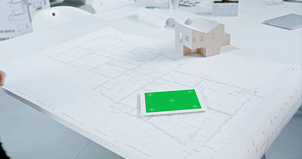 Workspace featuring a digital tablet with a green screen and a house model placed on architectural blueprints. The setting includes a clean, organized desk environment suitable for design and planning activities.