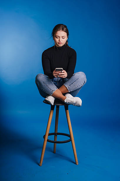 istock A young woman casually seated on a stool while holding her smartphone against a blue background 2242339887