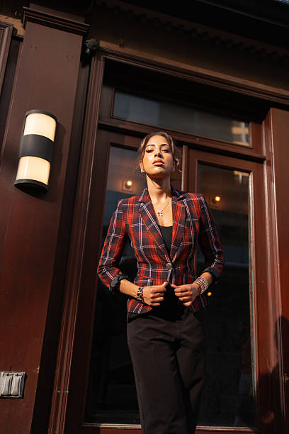 Confident young adult woman standing in an urban environment, wearing a stylish plaid blazer with sunlight on her face