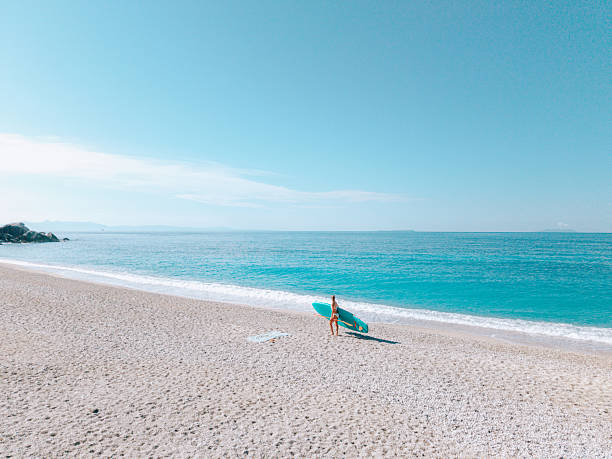 Woman in swimsuit standing on a quiet sandy beach holding a surfboard near the ocean. Solo surfer on a peaceful coastline with clear blue water. Beach lifestyle and outdoor recreation.