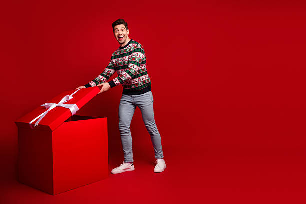 istock Funny Christmas moment as a handsome young man in a festive sweater pulls a giant red gift box against a bright red backdrop 2242352334