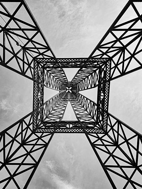 Abstract architectural perspective of a steel lattice tower shot from below in black and white. The geometric symmetry and repeating metal patterns create a striking minimalist composition emphasizing structure, engineering, and design aesthetics.