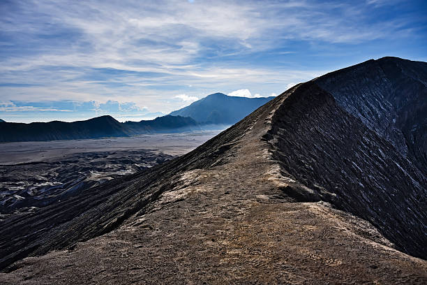 The crest of Mount Bromo offers an experience both striking and meditative. This narrow path, carved from volcanic ash, winds along the crater rim, overlooking the vast sand sea of ​​Bromo Tengger Semeru National Park. The air is thick with fine particles, the wind occasionally stirs up a cloud of dust, and each step echoes like a prayer to the power of nature.In the golden morning light, the barren and austere landscape reveals a raw, almost otherworldly beauty. The surrounding mountains loom on the horizon, bathed in a light mist, while the cracked earth bears witness to the volcano's ancient breath.Walking this fragile line between emptiness and sky is a sense of humility in the face of the vastness of the world. The landscape seems frozen in time, but the volcanic energy dormant beneath reminds us that nothing is ever completely calm here. A place of rare intensity, where every glance towards the horizon evokes the strength, solitude and wild beauty of Java.