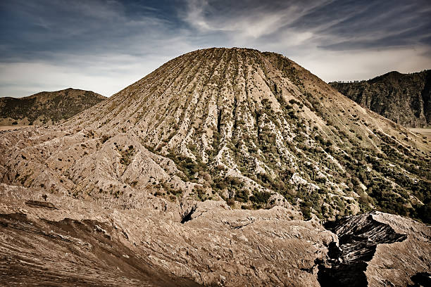 Towering over the Tengger caldera, Mount Batok rises like a perfect cone at the foot of the famous Mount Bromo, on the island of Java in Indonesia. Unlike its still-active neighbor, Mount Batok is an extinct volcano, covered in fine striations and sparse vegetation that highlight the rugged beauty of its slopes. The regular lines descending from its summit evoke a natural architecture, shaped by centuries of erosion, wind, and volcanic ash.In the golden morning light, its slopes are adorned with warm tones, oscillating between ochre and green, while the horizon reveals the surrounding volcanic hills. The contrast between the rough texture of the soil and the softness of the sky gives this landscape an almost spiritual dimension. Mount Batok is also a sacred place for the Tengger people, who consider it a protective mountain and participate in traditional ceremonies in the region. This iconic site in Bromo Tengger Semeru National Park embodies serenity amidst nature shaped by fire. A place of contemplation where one can feel both the strength and peace emanating from Indonesia's volcanoes.